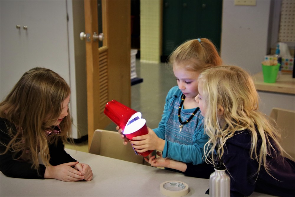 Three students performing a science experiment with two cups and a flashlight